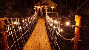 Un pont suspendu en bois, illuminé de guirlandes lumineuses, mène à une petite construction en bois.