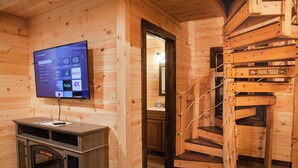 L’intérieur d’un chalet en bois est doté d’un téléviseur fixé au mur, qui diffuse des services de streaming, d’une cheminée et d’un escalier sur lequel est inscrit « SENTIERS DE RANDO ».