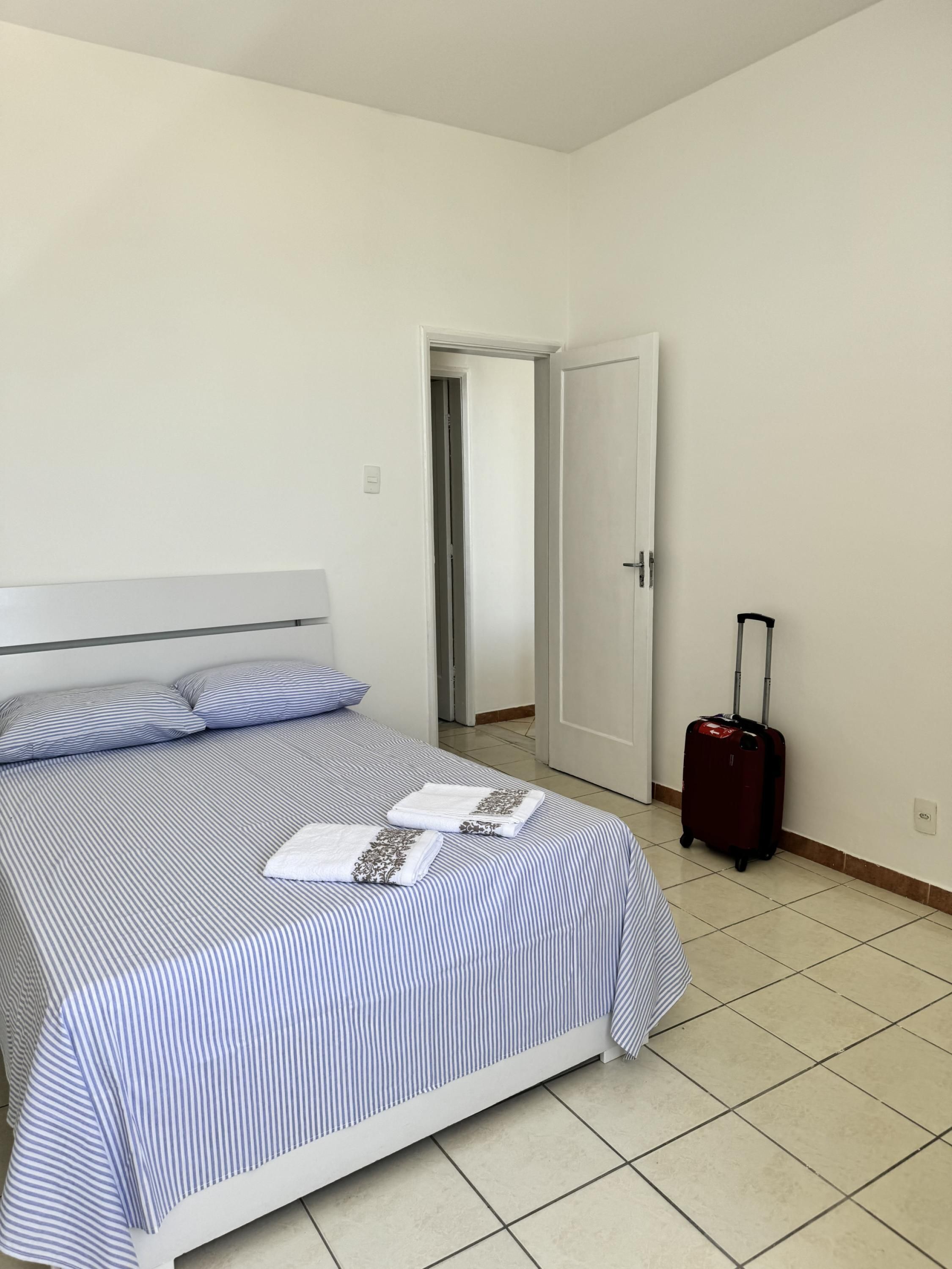 Een net opgemaakt bed met streepjesbeddengoed, een rode koffer en een gesloten deur in een eenvoudige kamer met witte muren en betegelde vloer.