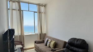 Un salón con sofá color beige, un sillón de cuero negro, una mesita y un televisor. Una ventana con cortinas ofrece vistas al mar.