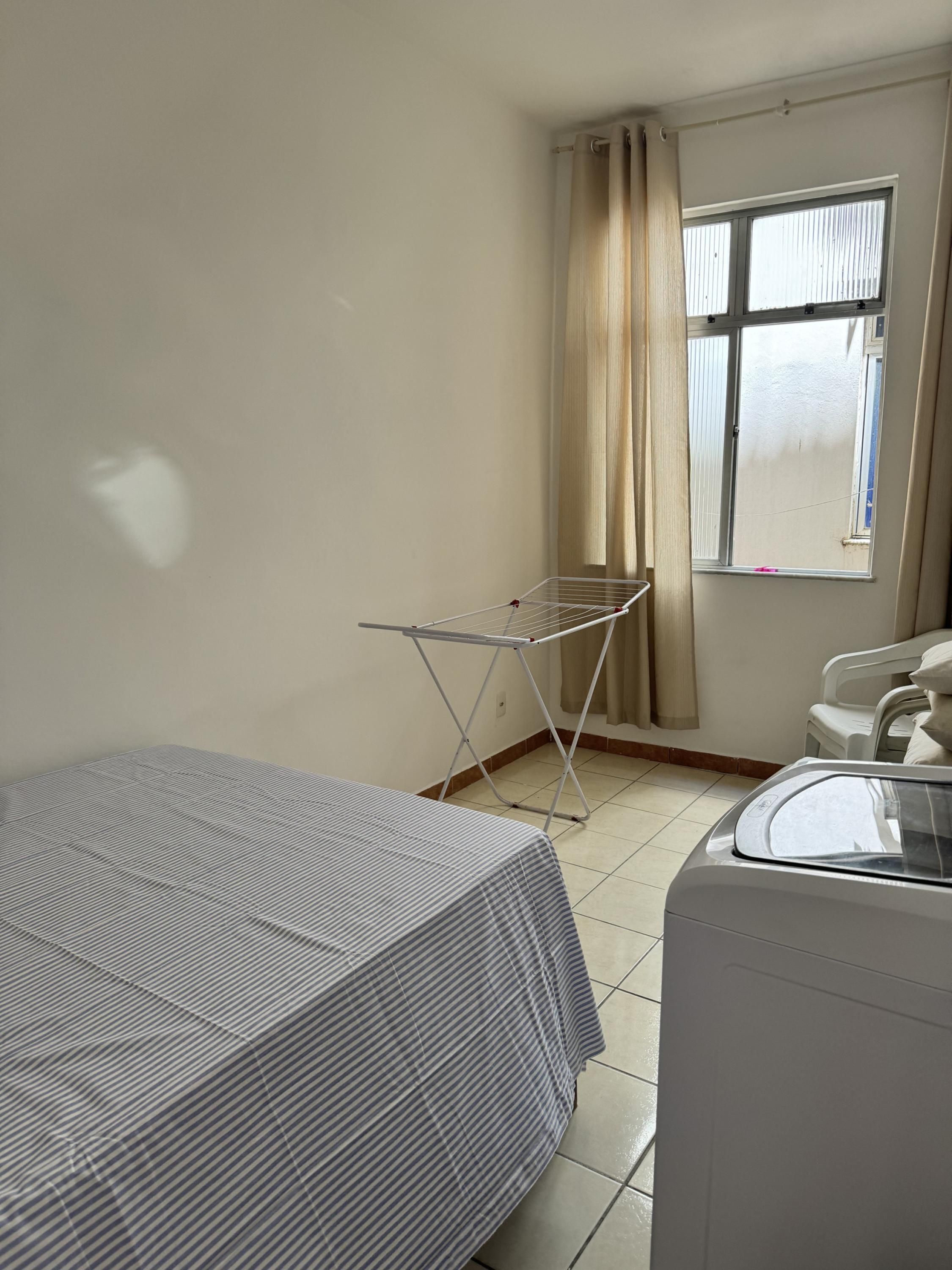 Una stanza piccola e pulita con un letto, uno stendino, una finestra con tende e una lavatrice.