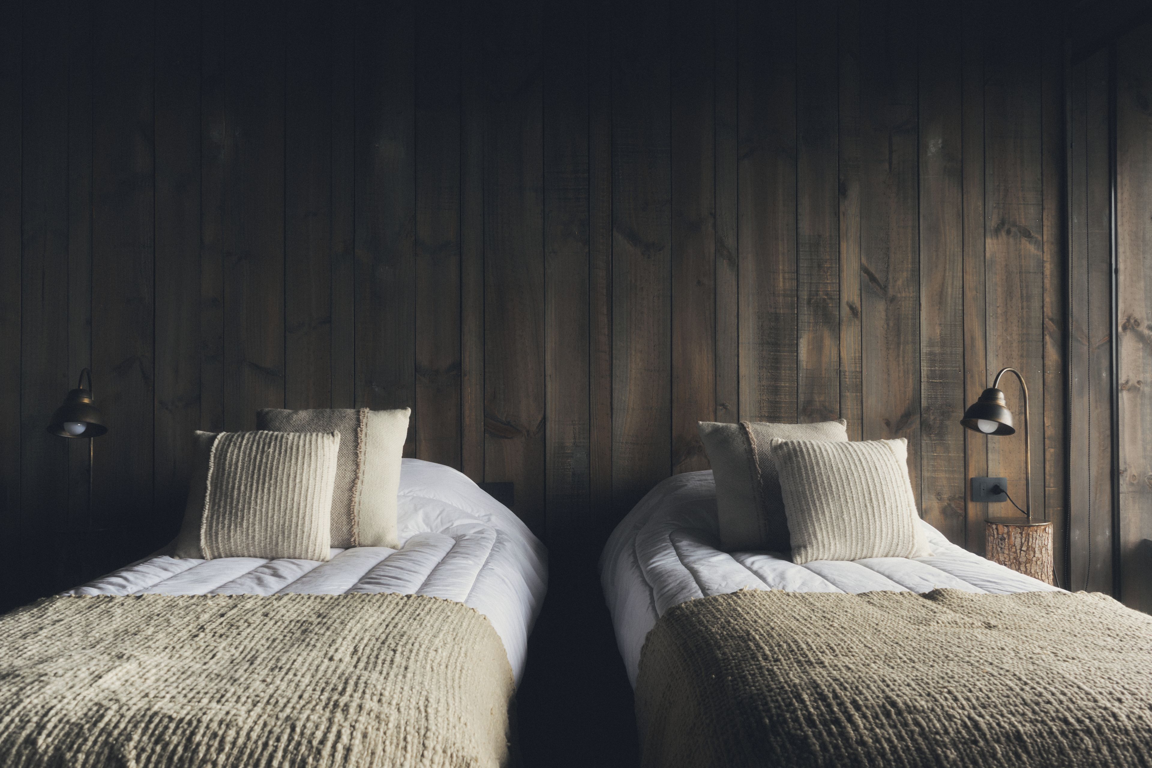 Twee enkele bedden met witte lakens en beige dekens, houten wanden en twee wandlampen.