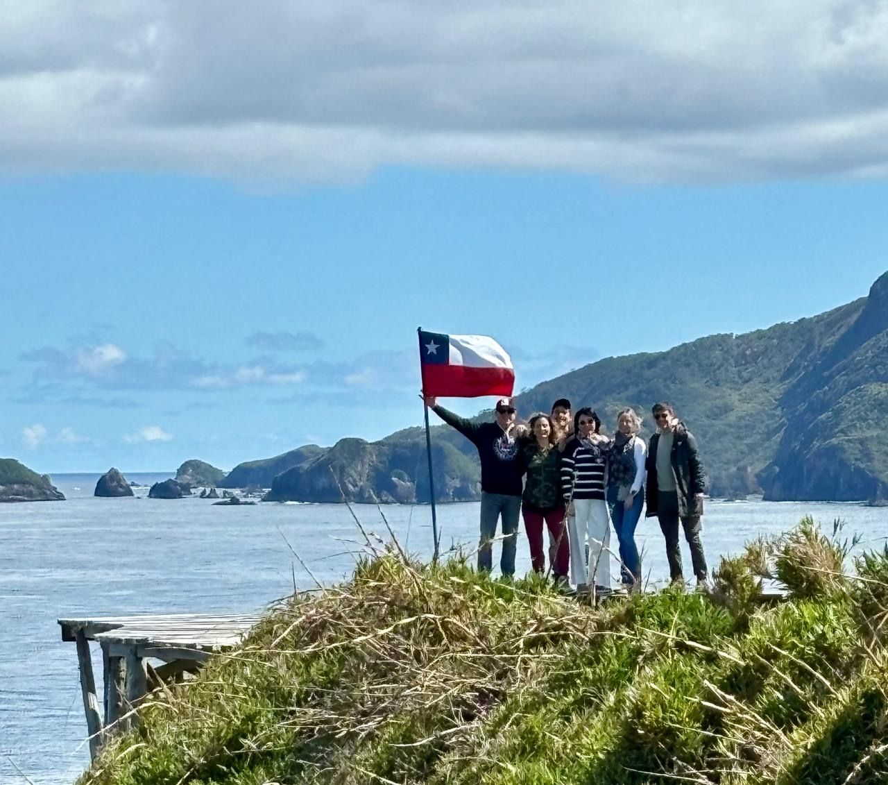 A group of people standing by a body of water with a flag, rocky terrain, and a clear sky.