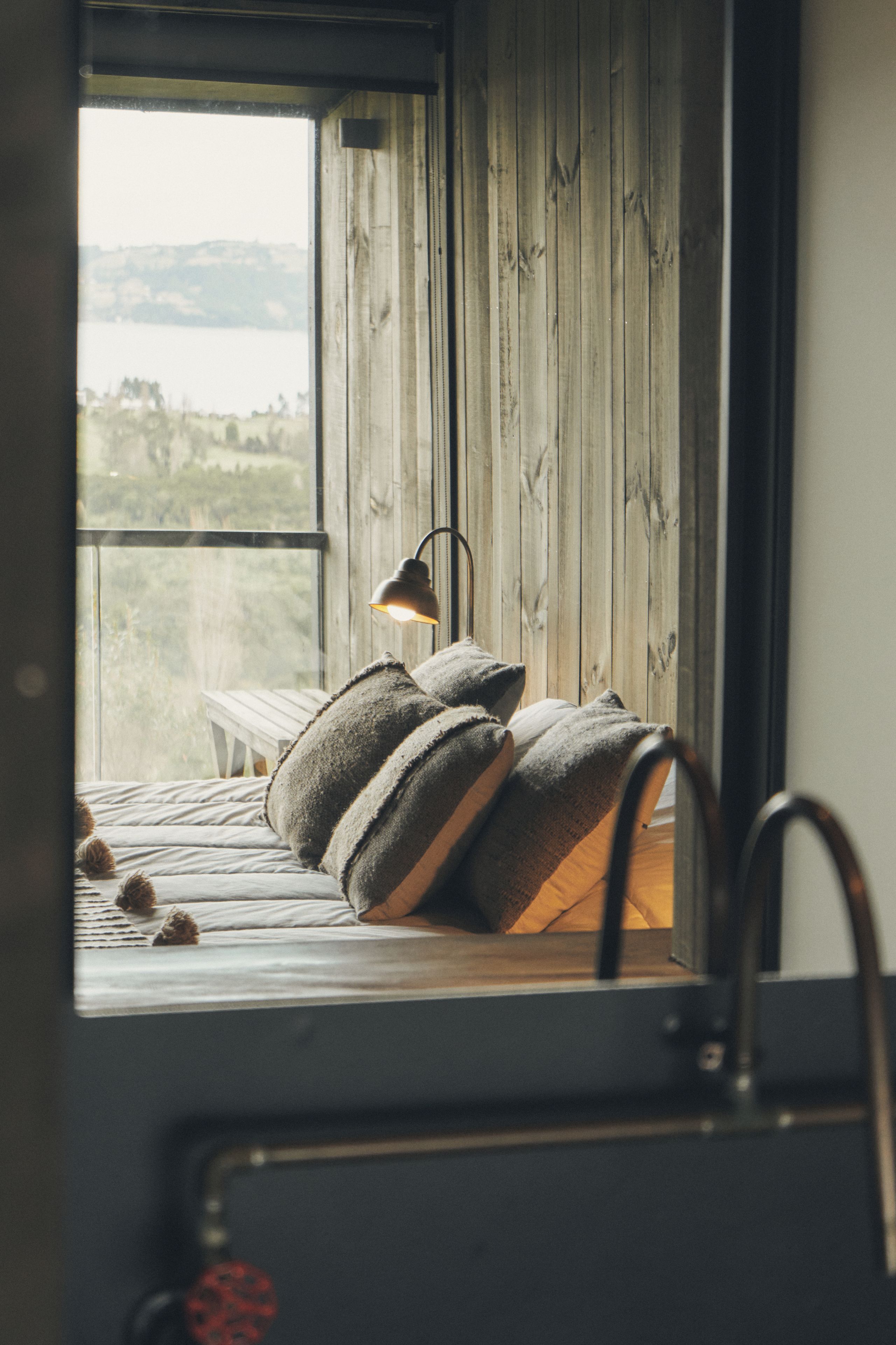 A bed with pillows and a lamp, a window with a view of the outdoors, and a wooden wall.