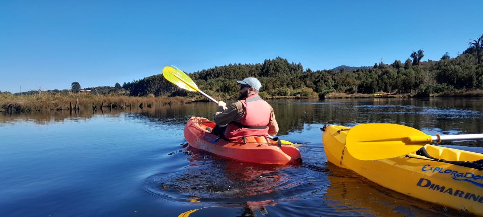 A person kayaking on a calm lake with a forested hillside in the background.