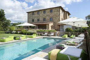A stone building with a swimming pool, green lounge chairs, and umbrellas.