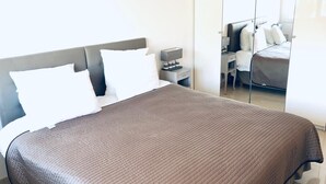 Een netjes opgemaakt bed met een bruine deken, een nachtkastje met een lamp en een garderobe met spiegel.