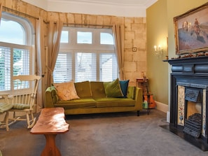 Un salón acogedor con un sofá verde, un sillón mecedor de madera, una mesita y una chimenea con un cuadro encima.