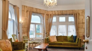 A room with a green sofa, a wooden coffee table, and a white chair. There are two windows with curtains, a chandelier, and a door with a brass handle.