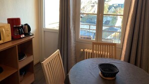 Una habitación con una mesa de comedor, una silla, una estantería con electrodomésticos y una ventana con cortinas.