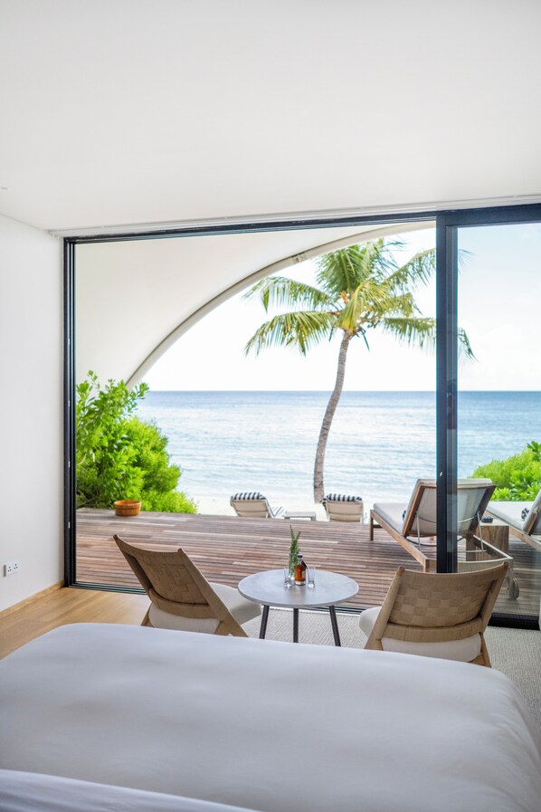 A bedroom with a large bed, a small round table, two chairs, and a view of the ocean through a glass door. - Silversands Beach House Adult only (St. George's)