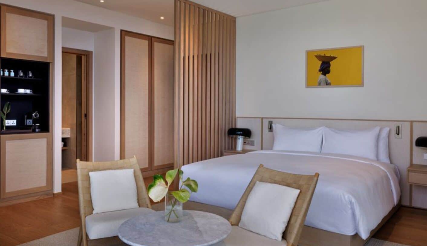 A hotel room with a bed, two chairs, a small table with a vase, a wooden cabinet, and a painting on the wall.