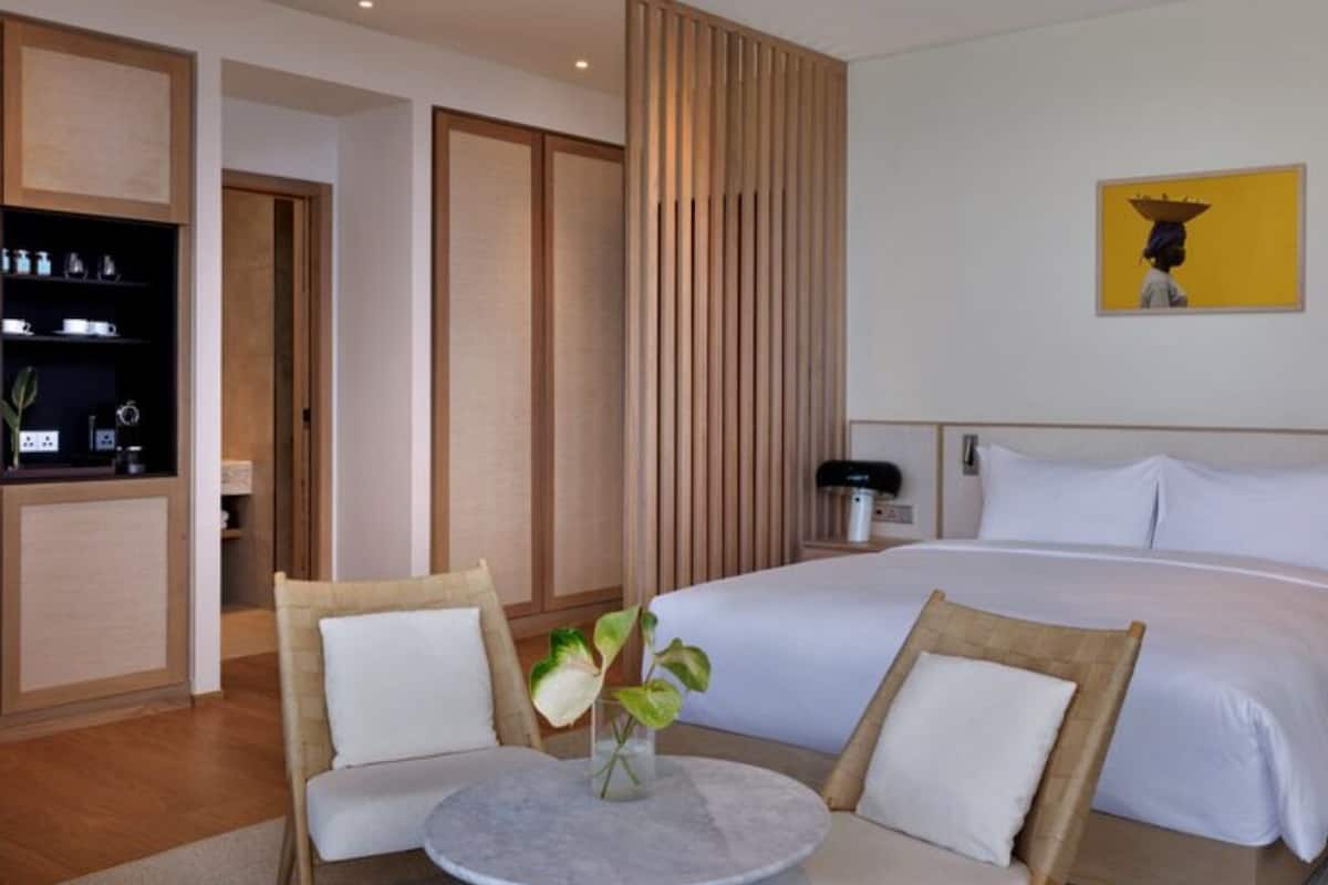 A hotel room with a bed, two chairs, a small table with a vase, a wooden cabinet, and a painting on the wall.