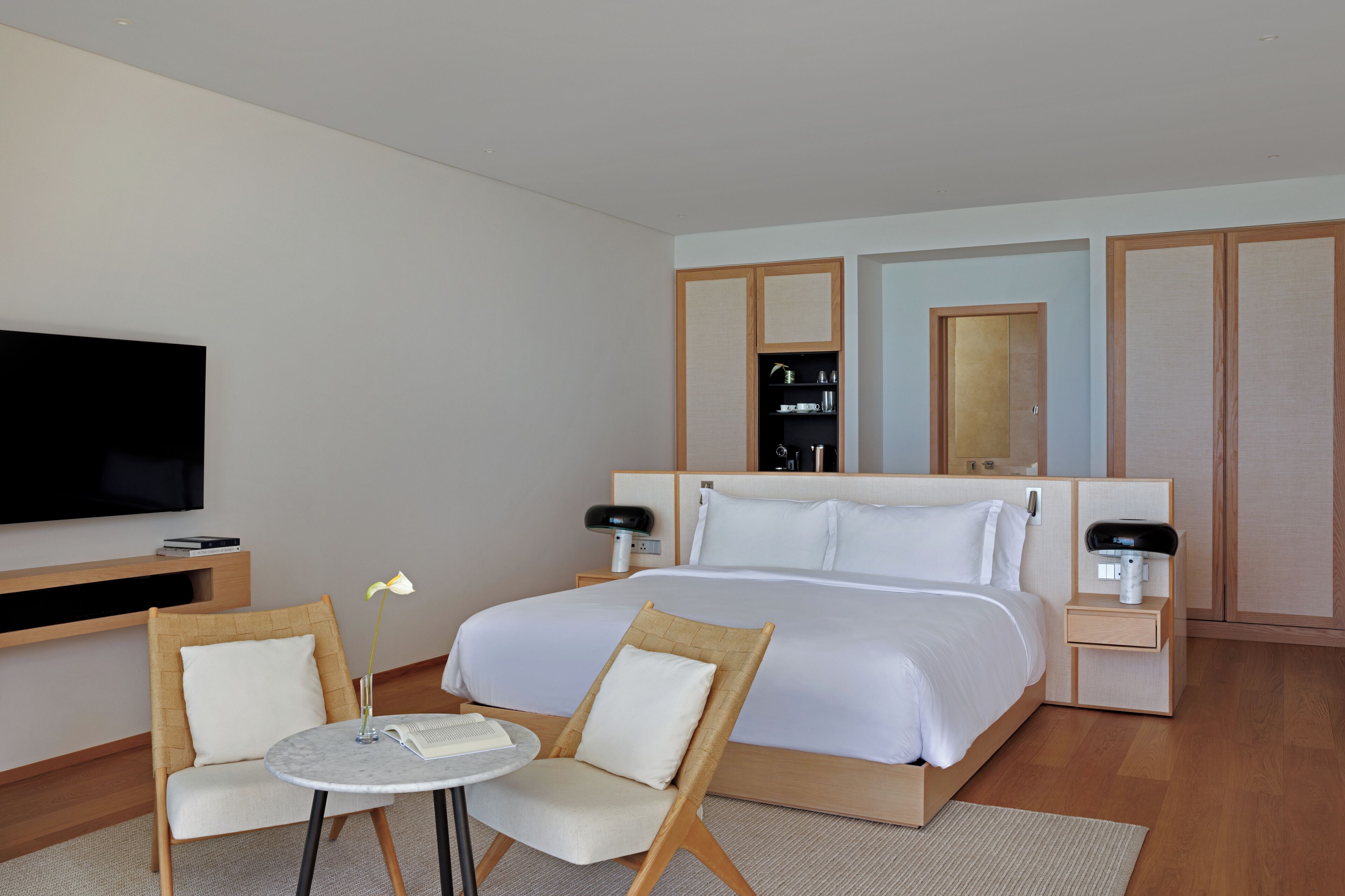 A hotel room with a large bed, a TV, a small table with chairs, and a bedside table with a lamp.
