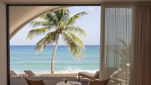 Habitación con una ventana grande que da a una playa, con una palmera, sillones de descanso y una cama.