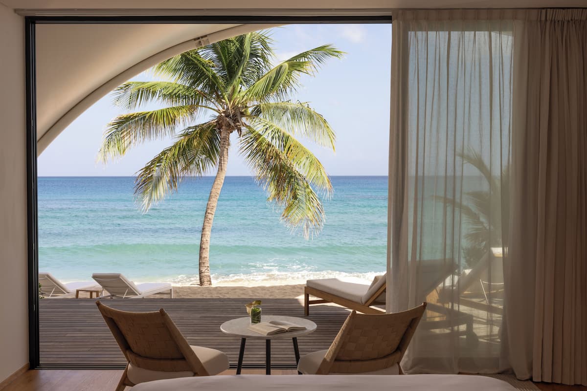 A room with a large window overlooking a beach, featuring a palm tree, lounge chairs, and a bed.
