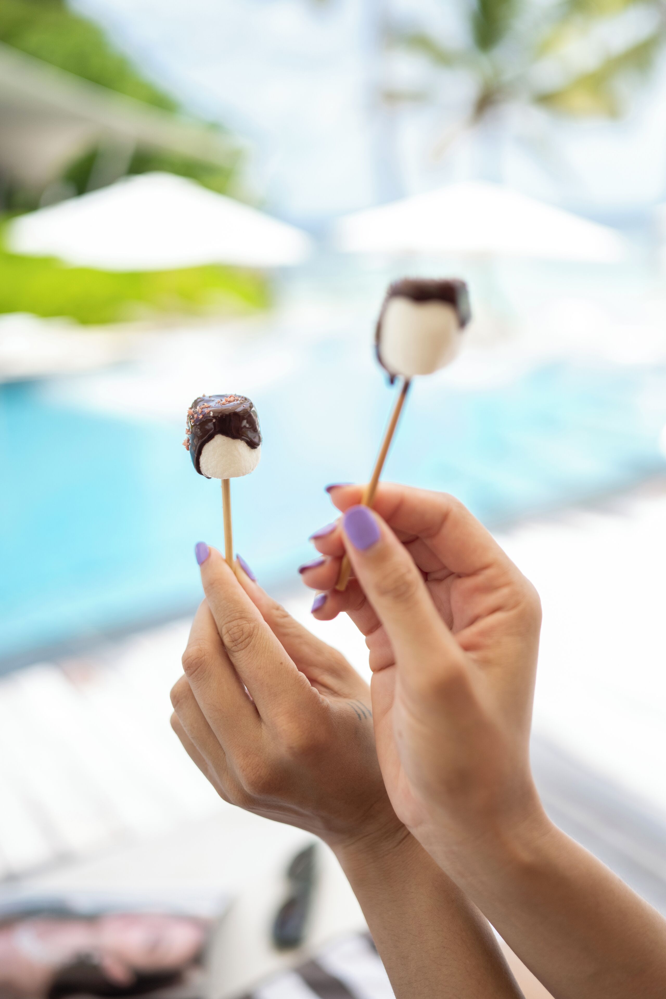 Two hands holding chocolate-covered marshmallow treats by a poolside.
