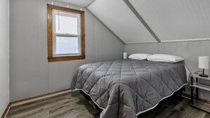 Un dormitorio con techo inclinado, una cama con colcha gris, una mesita de noche con lámpara y una ventana con persianas.