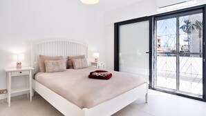 A modern bedroom with a white bed, two bedside tables with lamps, a large window with a view of buildings, and a sliding glass door. - Vilamoura Marina Villa  (Loulé)