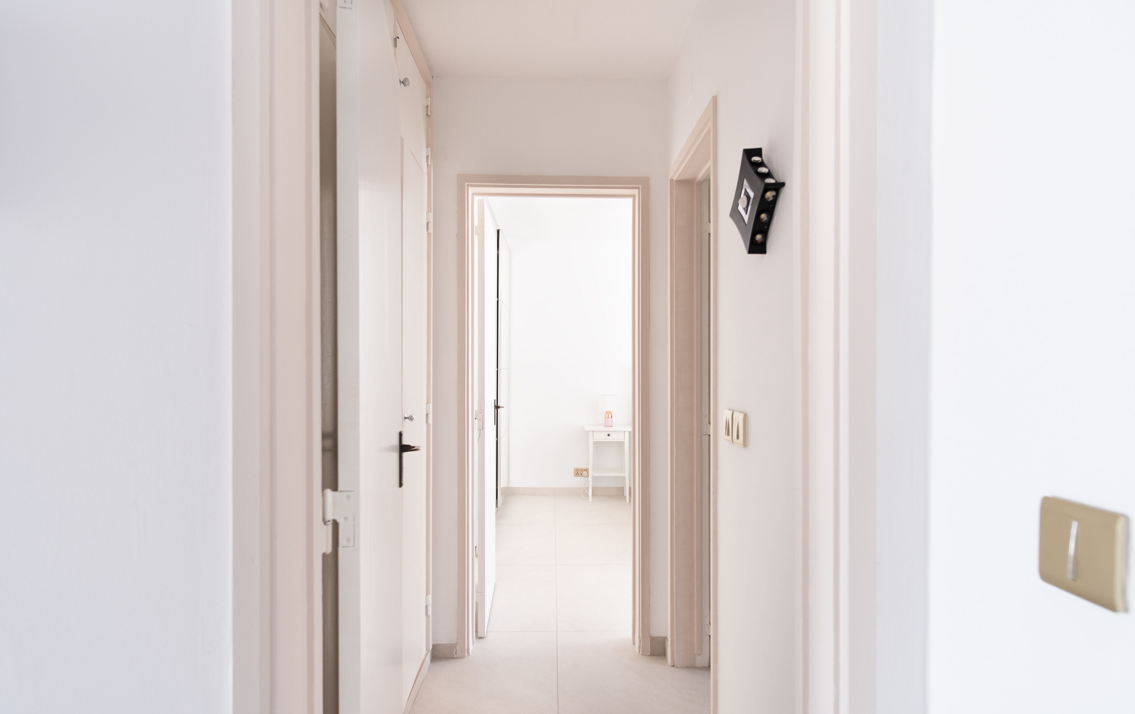 Un pasillo con paredes y puertas blancas que conduce a una habitación con una pequeña mesa blanca y un marco de fotos en la pared.
