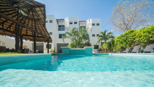 A swimming pool with clear blue water, surrounded by a modern building with balconies and a thatched roof structure. - Tranquilidad 3 Bedroom Home At El Cielo (Playa del Carmen)