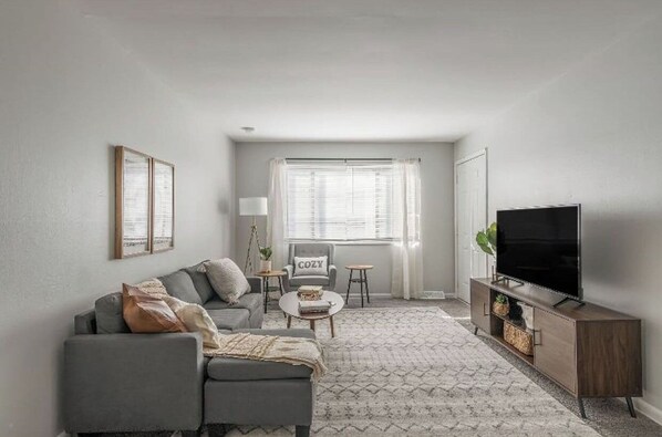 A living room with a grey sofa, a coffee table, a TV stand, and a window with curtains. - World Cup Home Base 30 Day Stay Netherlands Camp (Kansas City)