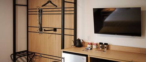 A compact room with a wooden wardrobe, a small refrigerator, a microwave, a coffee maker, and a TV on the wall.