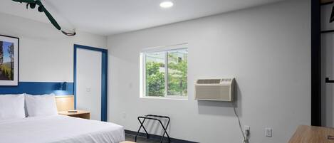 A modern hotel room with a bed, bedside table, bench, window, air conditioning unit, and a view of greenery outside.