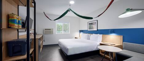 Une chambre d’hôtel comprenant un lit, un bureau, une chaise, une petite table, un canapé et un baldaquin vert suspendu.