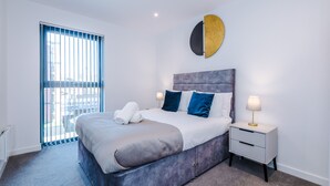 Un dormitorio moderno con una cama grande, una mesita de noche, una lámpara circular fijada a la pared y vistas a través de una ventana con persianas.