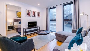 A modern living room with a sofa, a coffee table, and a TV. There is a window with curtains and a view of buildings outside. - Skeek Groundfloor Apartment with Wine Fridge (Birmingham)