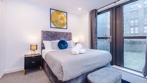 A modern bedroom with a large bed, a nightstand, a wall-mounted artwork, and a view of the city through a large window. - Skeek Groundfloor Apartment with Wine Fridge (Birmingham)