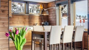 A wooden interior with a dining table and chairs, a vase of tulips, and a window with a view of a snowy landscape.