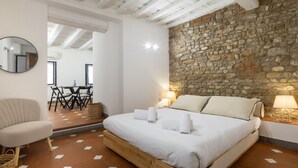 A bedroom with a stone wall, a bed with white linens, a chair, and a table with chairs. - San Giuseppe Apartment (Firenze)