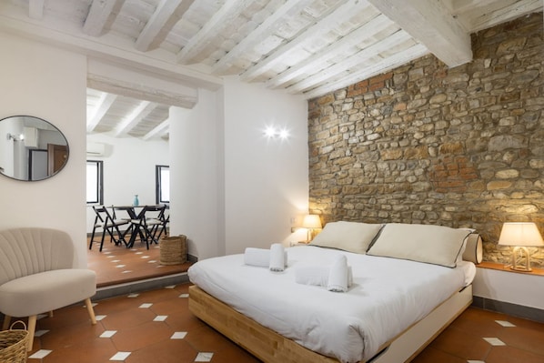 A bedroom with a stone wall, a bed with white linens, a chair, and a table with chairs.