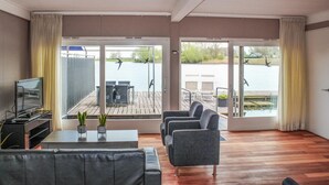 A modern living room with a wooden floor, grey sofa, and a TV on a stand. There are two large windows with curtains, offering a view of a lake and outdoor seating area. - Awesome ship in Lith with kitchen (Lith)