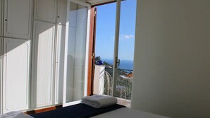 A room with a bed, a window with a view of the sea, and a balcony with a table and chairs.