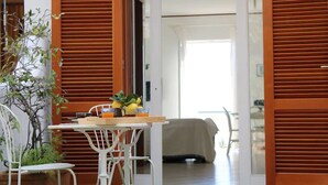 A room with a wooden door, a white chair, a small table with a laptop, a plant, and a bed covered with a white sheet.