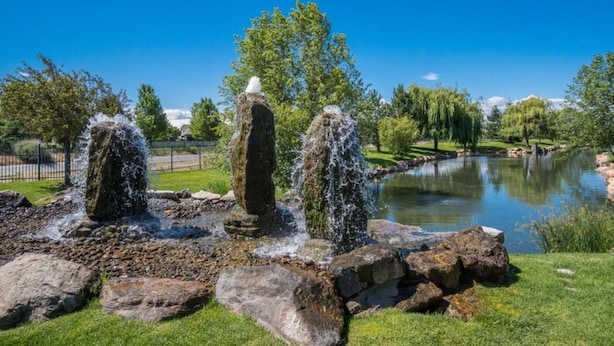 A tranquil garden with a pond, featuring a central rock formation with water spouts, surrounded by lush greenery and a clear blue sky. - Modern, king beds, pools, parks, pet OK, 5 mins toYMCA ,Store and top golf (Meridian)