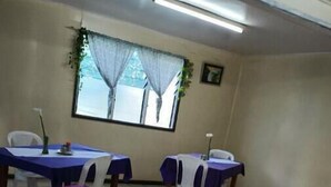 Une pièce avec quatre tables, chacune recouverte d’une nappe violette et entourée de chaises blanches. Il y a une fenêtre avec un rideau, une plante accrochée au mur et un écran de projection.