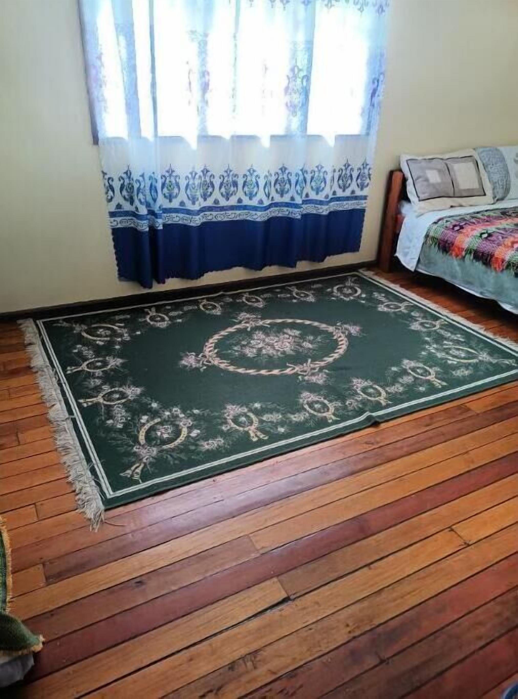 A room with a large green patterned rug, wooden flooring, a bed with a colorful blanket, and a window with patterned curtains.