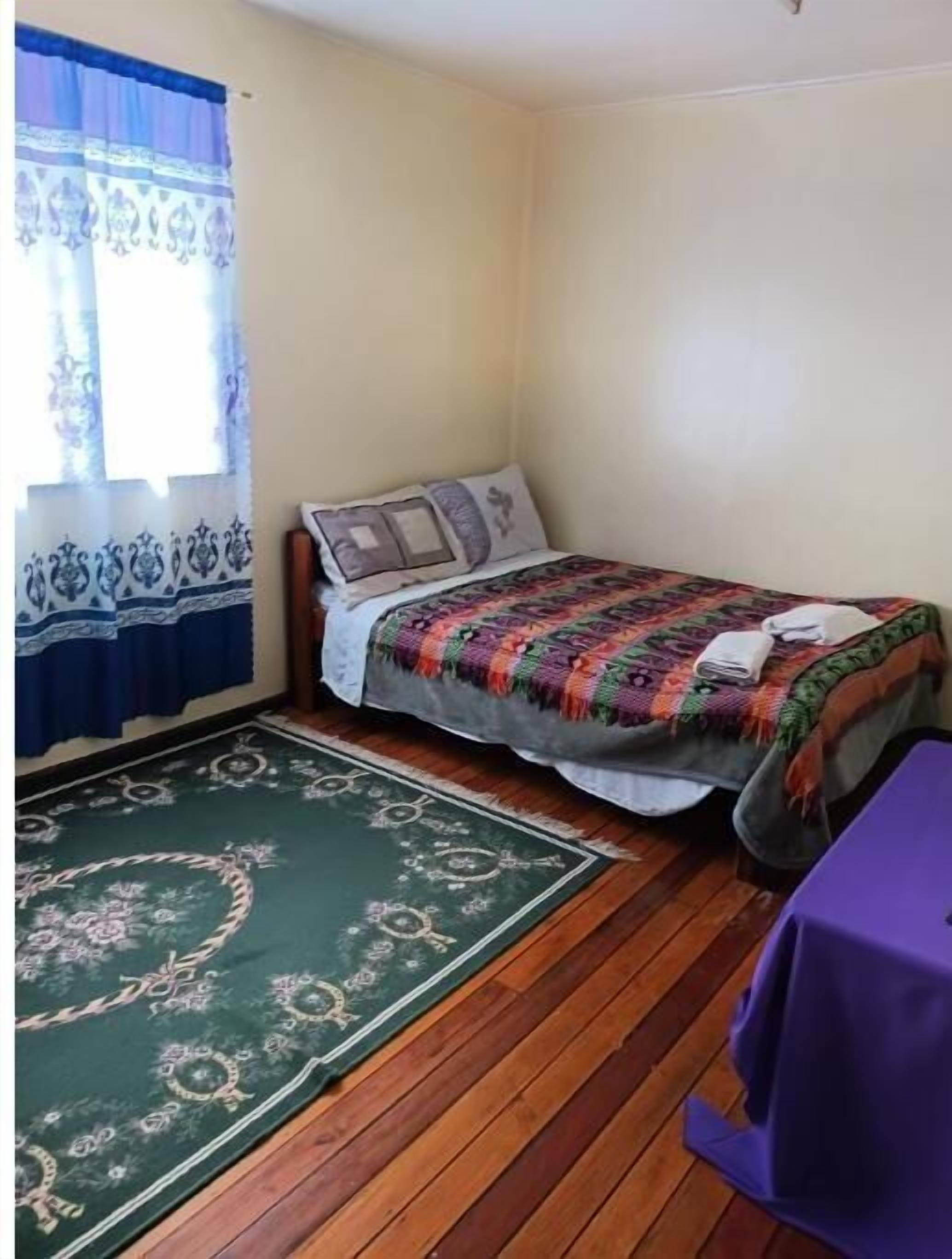 A small, single-bed room with a wooden floor, a green patterned rug, a purple table, and a window with blue curtains.