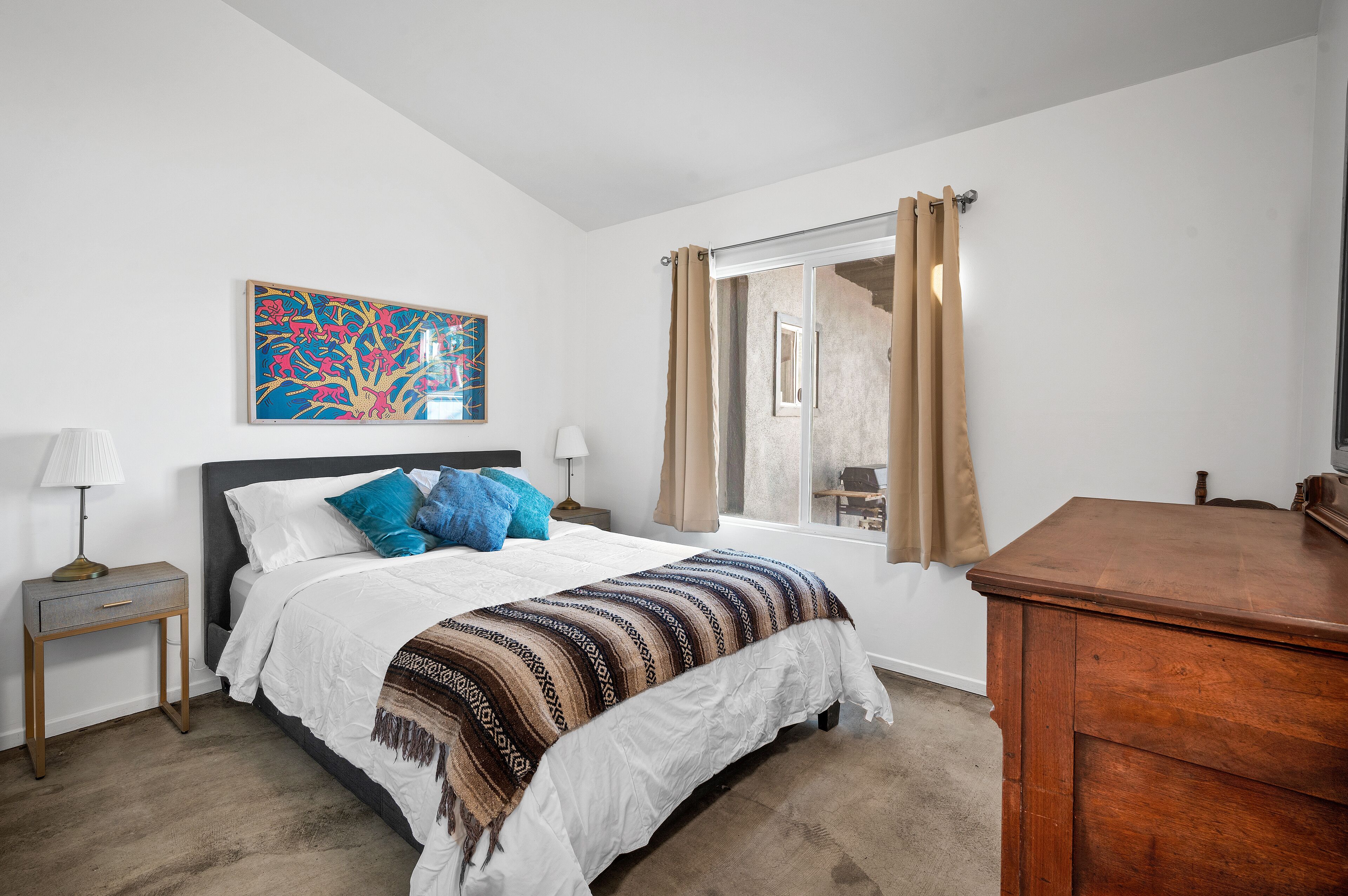 A bedroom with a bed, a nightstand, a wooden dresser, a window with curtains, and a colorful abstract painting on the wall.