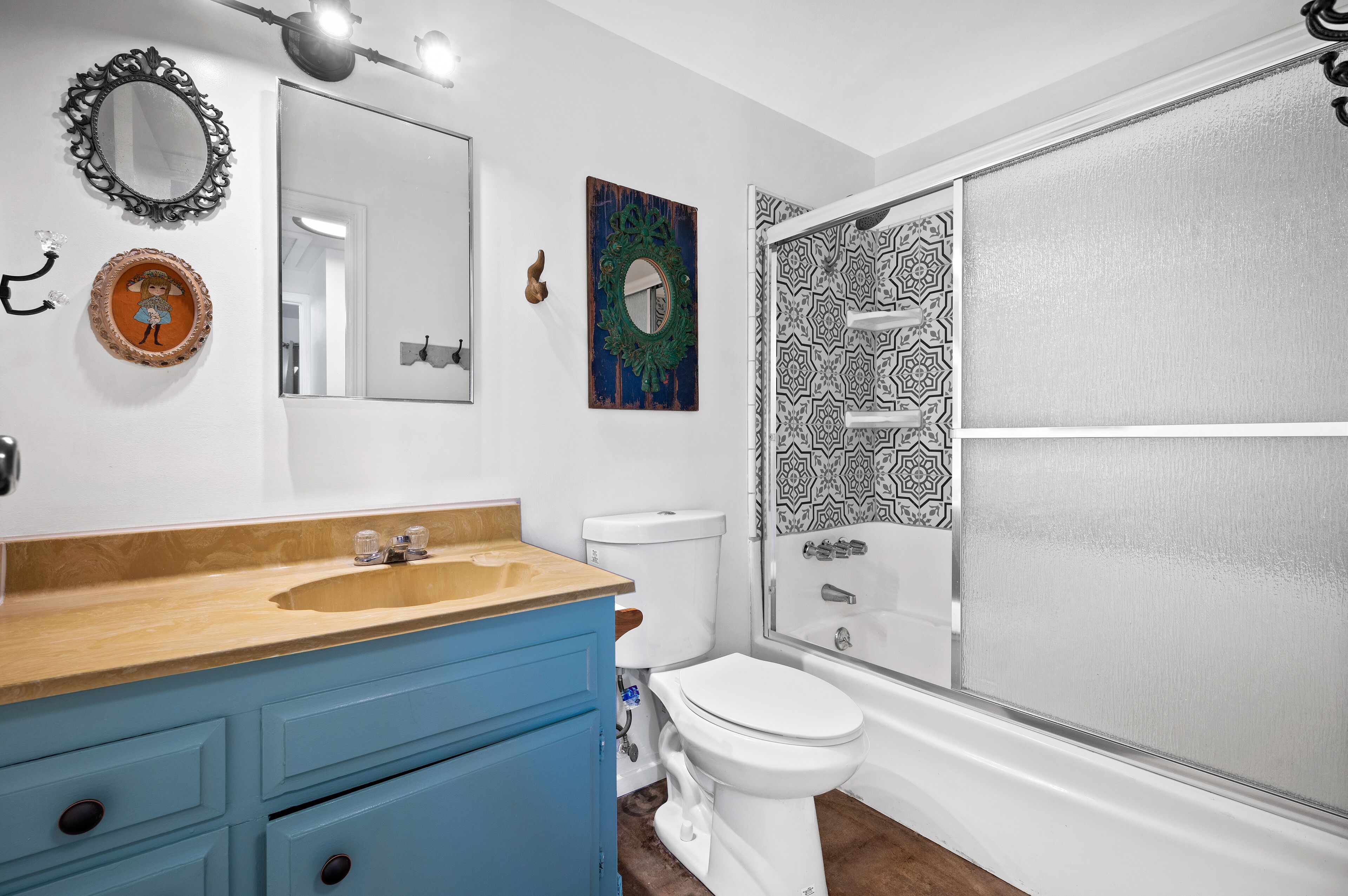 A bathroom with a wooden vanity, a white toilet, a shower with a patterned glass door, and decorative mirrors and wall art.