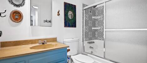 A bathroom with a wooden vanity, a white toilet, a shower with a patterned glass door, and decorative mirrors and wall art.