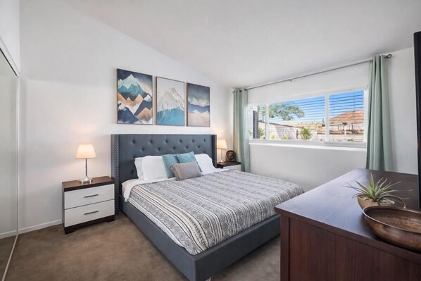 A modern bedroom with a large bed, a nightstand, a dresser, and a window with a view of buildings.