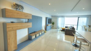 A modern living room with a wooden console, a blue herringbone wall, a sofa, a dining area with a white table and bar stools, and a large window with curtains. - Modern, large 3bedroom, spectacular views GOSLM-9 (Sliema)