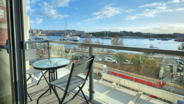 Un balcón con mesa y sillas, con vistas a una marina llena de barcos y un paisaje urbano lejano.