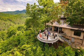Une terrasse circulaire en plein air, dotée d’une balustrade en pierre, entourée d’une végétation luxuriante et offrant une vue sur un paysage montagneux.