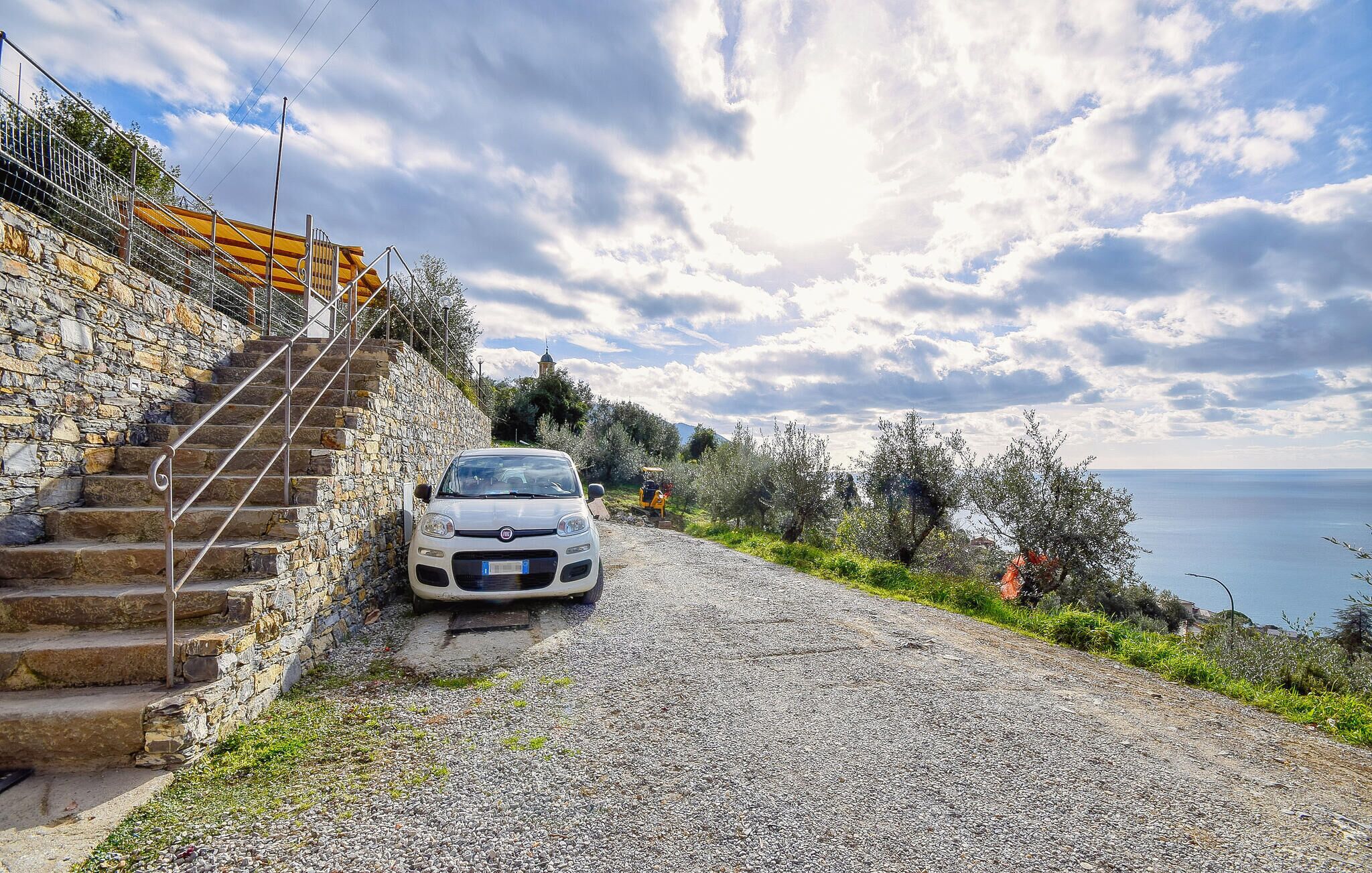 A white car parked on a gravel road beside a stone staircase leading to a building with a yellow awning.